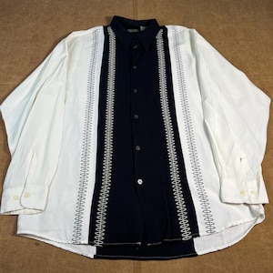 Puede incluir: Una camisa de manga larga con un panel central azul marino y paneles laterales blancos. El panel azul marino está adornado con bordados blancos verticales. La camisa tiene cuello y puños abotonados.