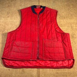 May include: A quilted red vest with a navy blue collar and a full-length zipper. The vest has two large front pockets and a quilted design throughout. The vest is sleeveless and appears to be made of a durable material.