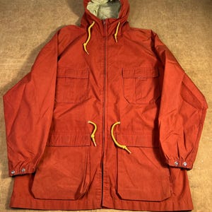 May include: A rust-colored hooded jacket with a drawstring hood and waist. The jacket has four front pockets, a zipper closure, and yellow drawstrings. The interior lining of the hood is a light green color. The sleeves have buttoned cuffs.