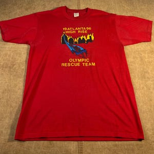May include: Red t-shirt with short sleeves. The shirt features a graphic with the text "19 ATLANTA 96 HIGH RISE" and "OLYMPIC RESCUE TEAM". The graphic includes a blue and red design with yellow accents.