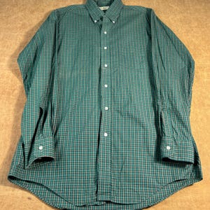 May include: A green and white checked button-down shirt with long sleeves. The shirt features a button-down collar and a chest pocket. The fabric has a subtle pattern of thin red lines. The shirt is made of a lightweight material.