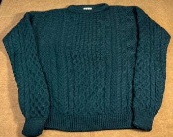 May include: A dark teal cable-knit sweater with a crew neck and ribbed cuffs and hem. The sweater features intricate cable patterns across the front and sleeves, offering a textured appearance. The garment is made of a thick, warm material.