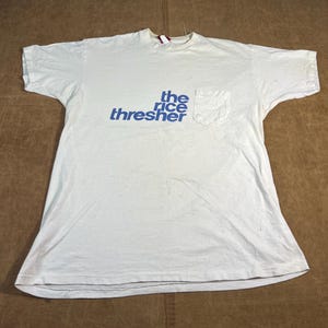 Può includere: T-shirt bianco con taschino e scritta blu "the rice thresher". La maglietta ha girocollo e maniche corte. Il tessuto appare leggermente usurato.