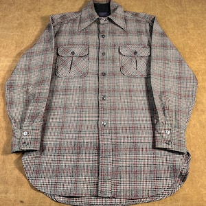 May include: A long-sleeved button-down shirt with a plaid pattern in shades of gray, red, and black. The shirt has two chest pockets with button closures and a classic collar. The fabric appears to be a woven material.