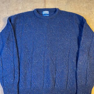 May include: A navy blue crew neck sweater with a textured diamond pattern. The sweater has flecks of red, white, and yellow throughout the fabric. The cuffs and hem are ribbed. A small label is visible at the neckline.