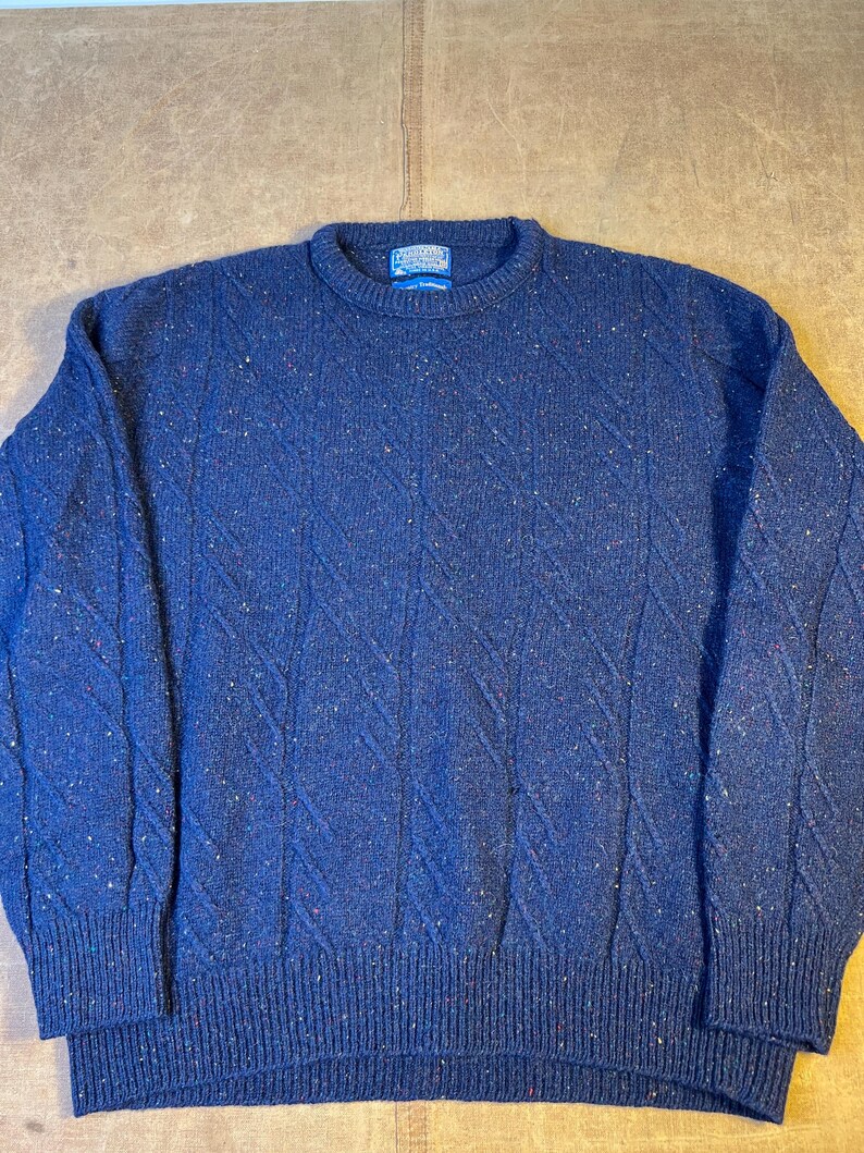 May include: A navy blue crew neck sweater with a textured diamond pattern. The sweater has flecks of red, white, and yellow throughout the fabric. The cuffs and hem are ribbed. A small label is visible at the neckline.