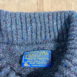 May include: Close-up of a gray wool sweater with flecks of color. The blue label reads "PENDLETON WOOLEN MILLS" and "100% VIRGIN WOOL" with "MADE IN U.S.A." The sweater is a size L.