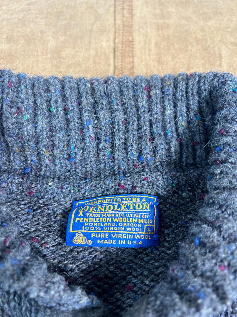 May include: Close-up of a gray wool sweater with flecks of color. The blue label reads "PENDLETON WOOLEN MILLS" and "100% VIRGIN WOOL" with "MADE IN U.S.A." The sweater is a size L.