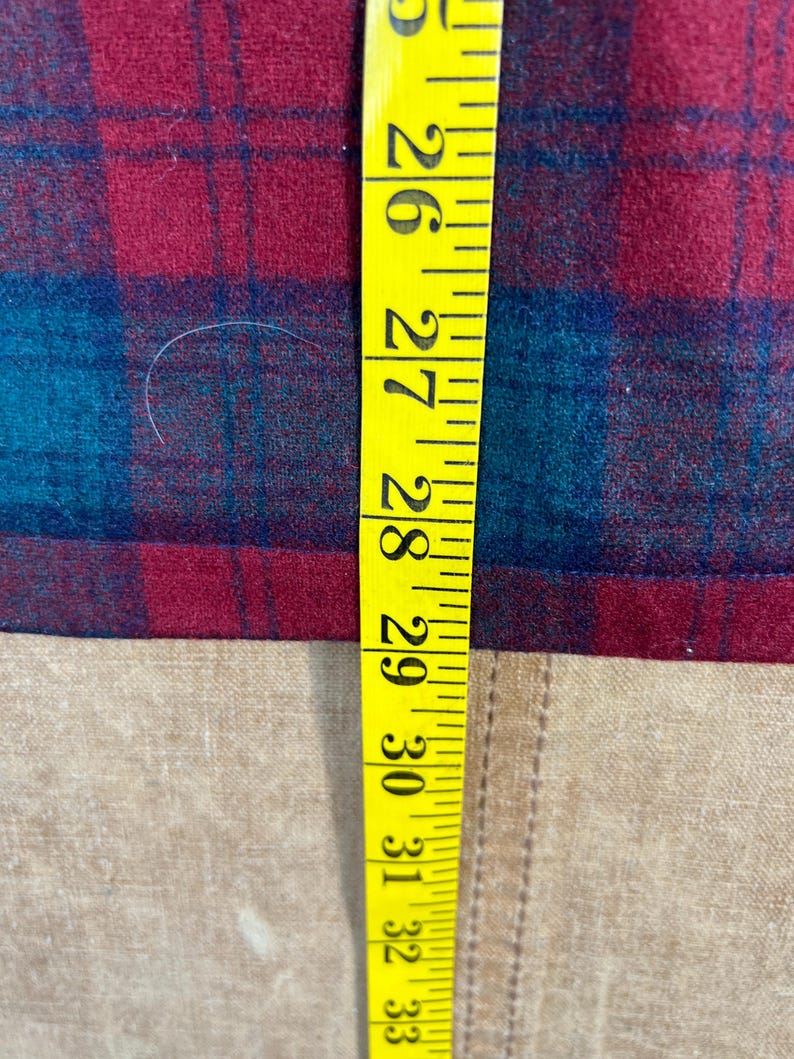 May include: A yellow measuring tape shows measurements from 66 to 84 cm. The tape is next to a red and teal plaid fabric. The fabric is on a tan surface.