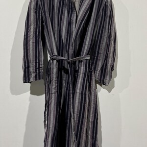May include: A striped robe in shades of gray and black hangs on a wooden hanger. The robe features vertical stripes and a matching belt. The garment is made of a lightweight fabric, suitable for lounging or as a cover-up.