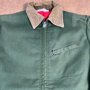 May include: Green work jacket with a brown corduroy collar and a red lining. The jacket has a zipper closure and a small pocket on the chest. The jacket is made of a durable fabric.
