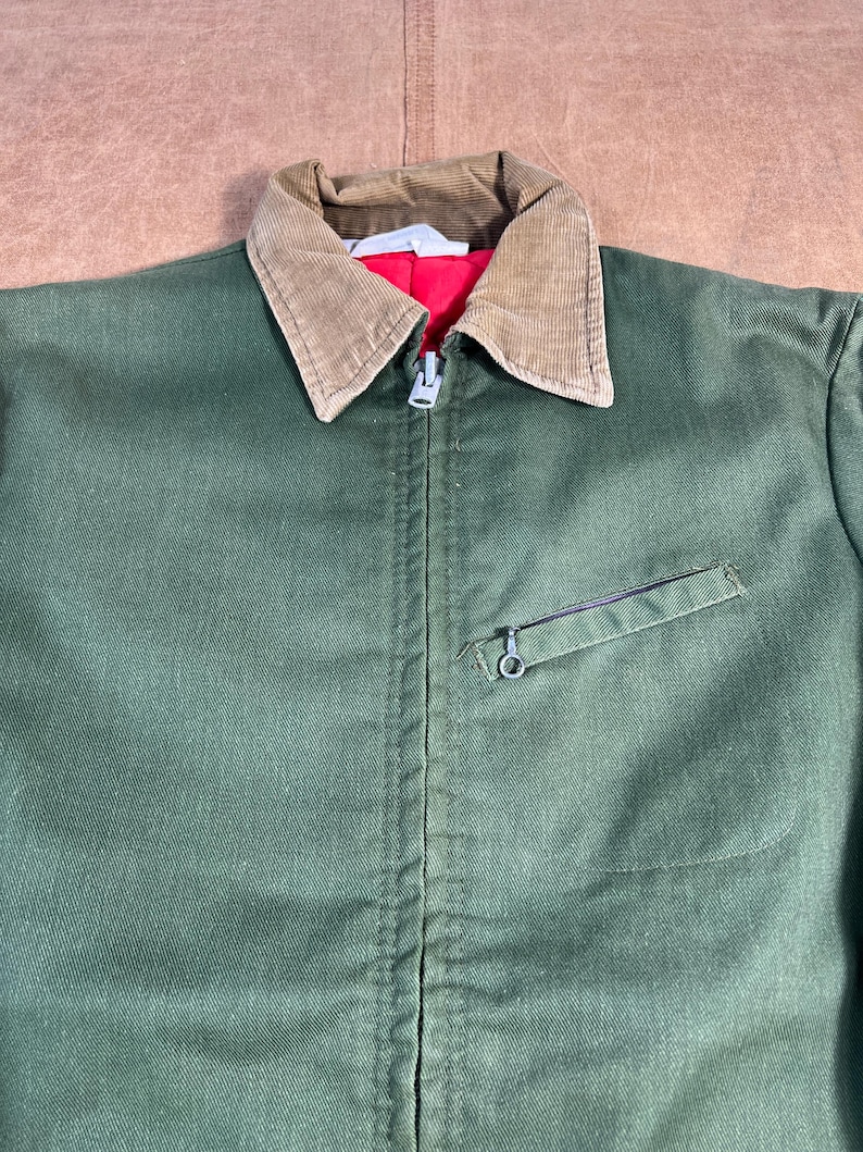 May include: Green work jacket with a brown corduroy collar and a red lining. The jacket has a zipper closure and a small pocket on the chest. The jacket is made of a durable fabric.