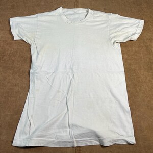 Peut inclure: Un t-shirt blanc uni à manches courtes, posé à plat sur une surface marron. Le t-shirt a un col rond et semble être fait d'un tissu doux et léger. Le t-shirt est légèrement froissé.