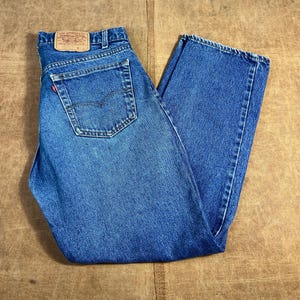 Puede incluir: Vaqueros azules de mezclilla con un diseño clásico de cinco bolsillos. Los vaqueros tienen una etiqueta roja de Levi's en el bolsillo trasero y un parche de cuero en la cintura. Los vaqueros están doblados y colocados sobre una superficie marrón.