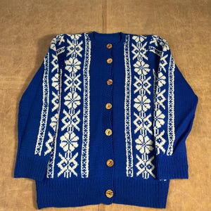 May include: A blue cardigan sweater with a cream-colored geometric pattern. The sweater has a round neck, long sleeves, and wooden buttons down the front. The pattern is repeated on the sleeves and front panels.