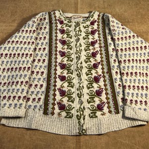 Puede incluir: Un cárdigan crema con botones y diseño floral. El suéter presenta un patrón repetido de pequeñas flores azules y granates en las mangas y un panel central con motivos florales granates y verdes más grandes. El cuello tiene una pequeña etiqueta "Woolrich".