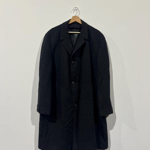 Peut inclure: Un pardessus noir charbon de bois avec un col cranté et trois boutons, suspendu à un cintre en bois. Le manteau est contre un mur blanc. Le tissu du manteau semble être texturé.