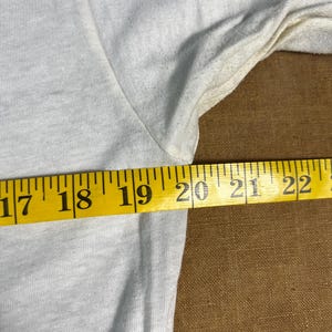 May include: Close-up of a light gray t-shirt with a yellow measuring tape across the fabric. The tape reads from 17 to 22 inches. The shirt's fabric is slightly wrinkled.