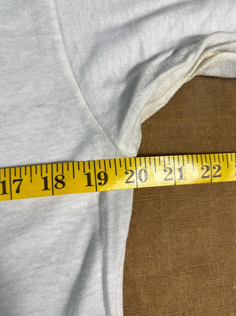 May include: Close-up of a light gray t-shirt with a yellow measuring tape across the fabric. The tape reads from 17 to 22 inches. The shirt's fabric is slightly wrinkled.