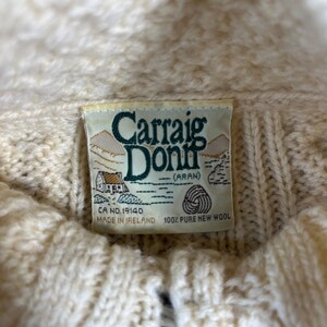 May include: Close-up of a cream-colored Aran sweater label. The label reads "Carraig Donn (Aran)" with a small illustration of a house and a wool symbol. The sweater is made of 100% pure new wool.