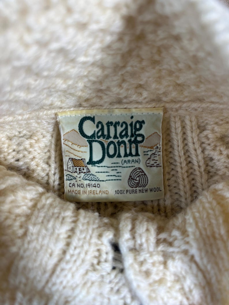 May include: Close-up of a cream-colored Aran sweater label. The label reads "Carraig Donn (Aran)" with a small illustration of a house and a wool symbol. The sweater is made of 100% pure new wool.