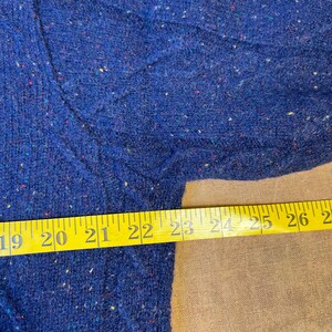 May include: Close-up of a blue sweater with a textured knit pattern. The sweater has flecks of various colors throughout the fabric. A yellow measuring tape is laid across the sweater, showing measurements from 19 to 26 inches.