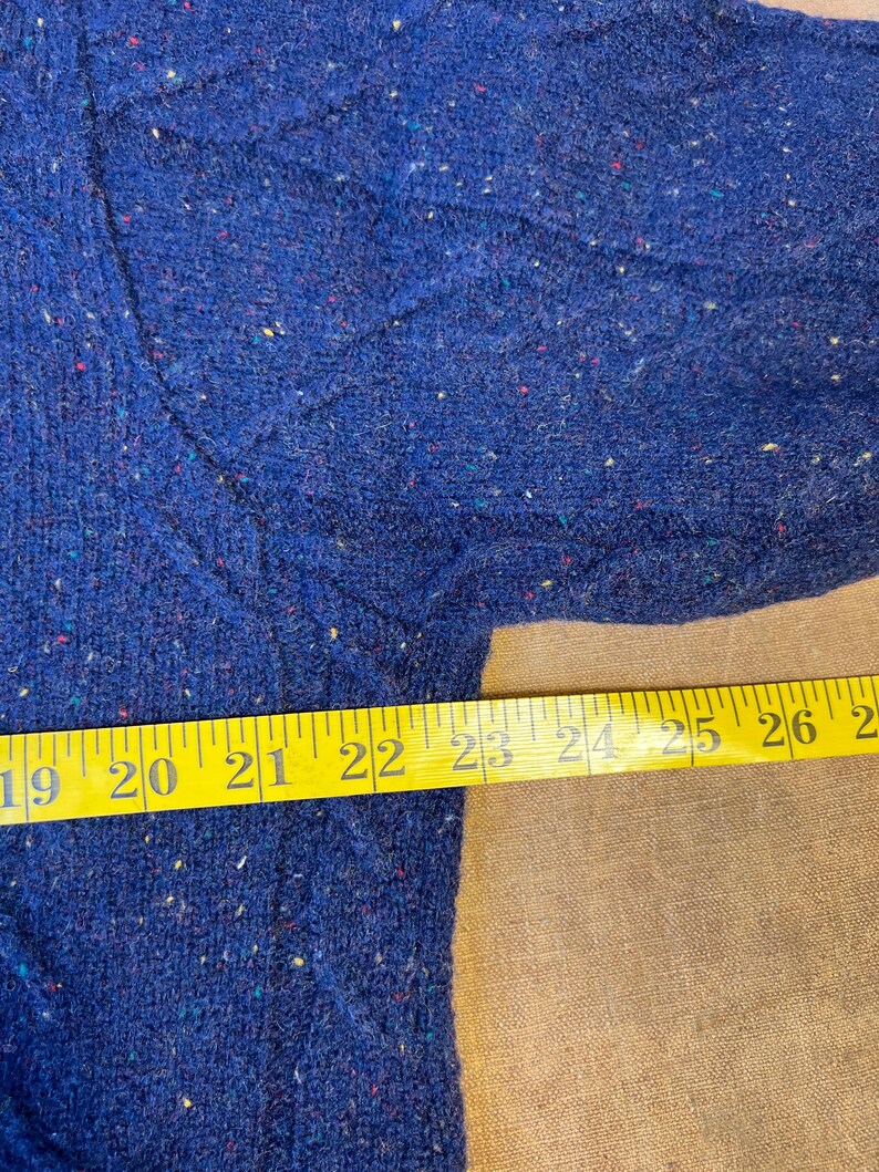 May include: Close-up of a blue sweater with a textured knit pattern. The sweater has flecks of various colors throughout the fabric. A yellow measuring tape is laid across the sweater, showing measurements from 19 to 26 inches.