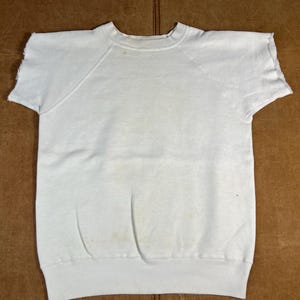 May include: A vintage white short-sleeved sweatshirt with a crew neck and raglan sleeves. The fabric shows signs of wear and discolouration. The bottom hem and sleeve edges have a raw, unfinished look.
