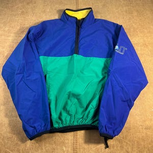 May include: A color-blocked pullover jacket in blue, green, and blue. The jacket has a yellow collar, a black zipper, and a small logo on the right sleeve. The jacket is made of a lightweight material and has elastic cuffs and hem.