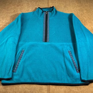 May include: Teal fleece pullover with a black zipper and trim. The collar is a mock neck style. The zipper and pocket edges have a decorative blue and black pattern. The long-sleeved top has two front pockets.