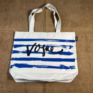 May include: White tote bag with blue horizontal brushstroke stripes. The word "Vogue" is written in black cursive across the front. The bag has two white handles and a small black tag that says "Vogue".