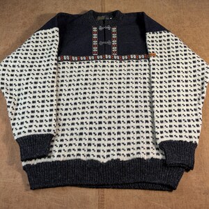 May include: A long-sleeve knit sweater featuring a dark charcoal upper section and a white lower body with a repeating black geometric pattern. The chest area includes a decorative placket with metal clasps and a colorful horizontal band.