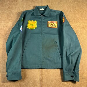 May include: A dark teal jacket with a zip, two front pockets, and buttoned cuffs. The jacket has several patches, including one with a bicycle and text that reads "Sparta-Elroy Bike & Rama". Another patch displays a deer.
