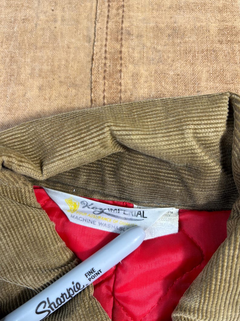 May include: Close-up of a tan corduroy jacket collar with a red quilted lining. The jacket's label reads "Key Imperial" and "Machine Washable". A silver Sharpie pen is in the foreground.
