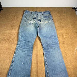 May include: Faded blue denim jeans with a flared leg design. The jeans show signs of wear and tear, including patches on the back pockets and distressed areas. The jeans are laid flat on a brown surface.