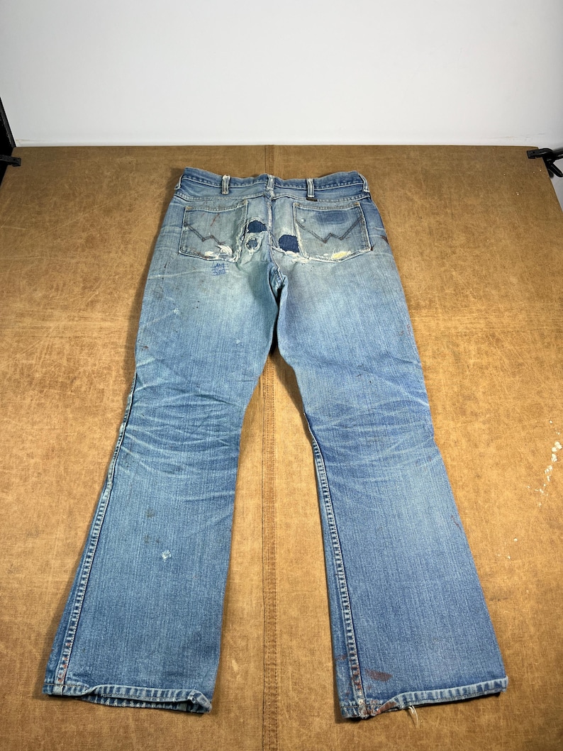 May include: Faded blue denim jeans with a flared leg design. The jeans show signs of wear and tear, including patches on the back pockets and distressed areas. The jeans are laid flat on a brown surface.