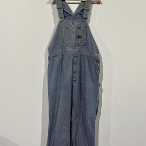 May include: Blue and white striped bib overalls hanging on a wooden hanger. The overalls have adjustable straps, a front bib pocket, and multiple pockets on the legs. The fabric has a vertical stripe pattern. The overalls are made of a durable material.