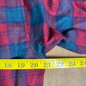 May include: Close-up of a red and teal plaid fabric. The fabric is measured with a yellow tape measure, showing measurements from 45.7 to 61 cm. The plaid pattern is a classic design.