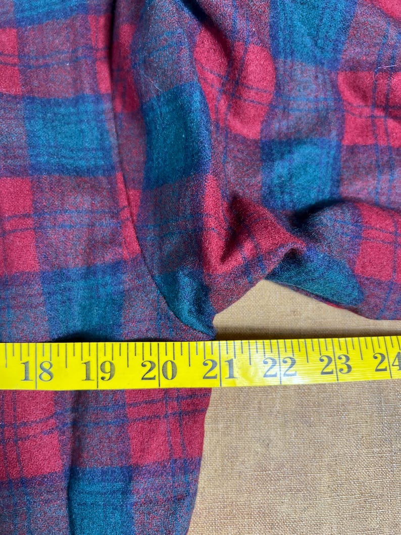 May include: Close-up of a red and teal plaid fabric. The fabric is measured with a yellow tape measure, showing measurements from 45.7 to 61 cm. The plaid pattern is a classic design.