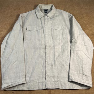 May include: A light gray jacket with a collared neckline and two chest pockets. The jacket has a button-up front and long sleeves. The fabric appears to be a textured linen or cotton blend. The jacket is laid flat on a brown surface.