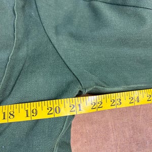May include: Close-up of a dark green garment with a yellow measuring tape. The tape shows measurements from 18 to 25 inches. The fabric has a textured appearance.