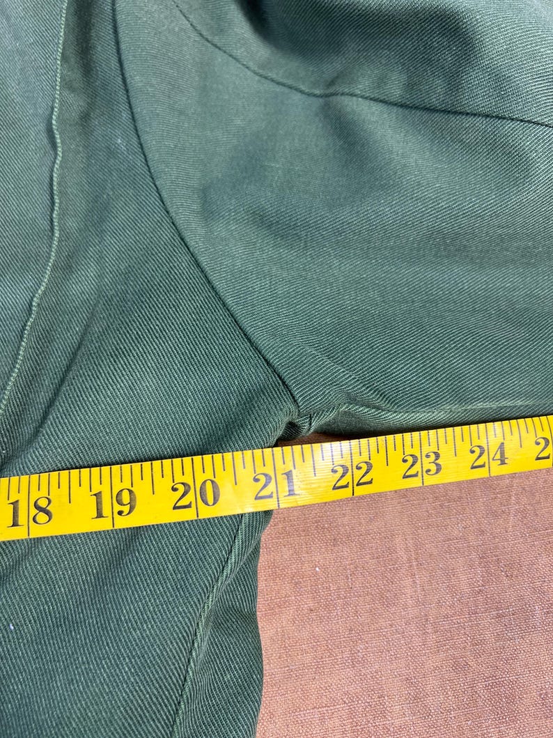 May include: Close-up of a dark green garment with a yellow measuring tape. The tape shows measurements from 18 to 25 inches. The fabric has a textured appearance.