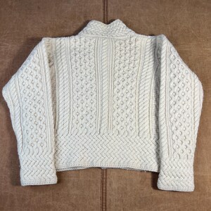 May include: Cream-colored, textured knit sweater with a high collar. The sweater features intricate cable knit and diamond patterns across the body and sleeves. The garment is laid flat on a brown leather surface.