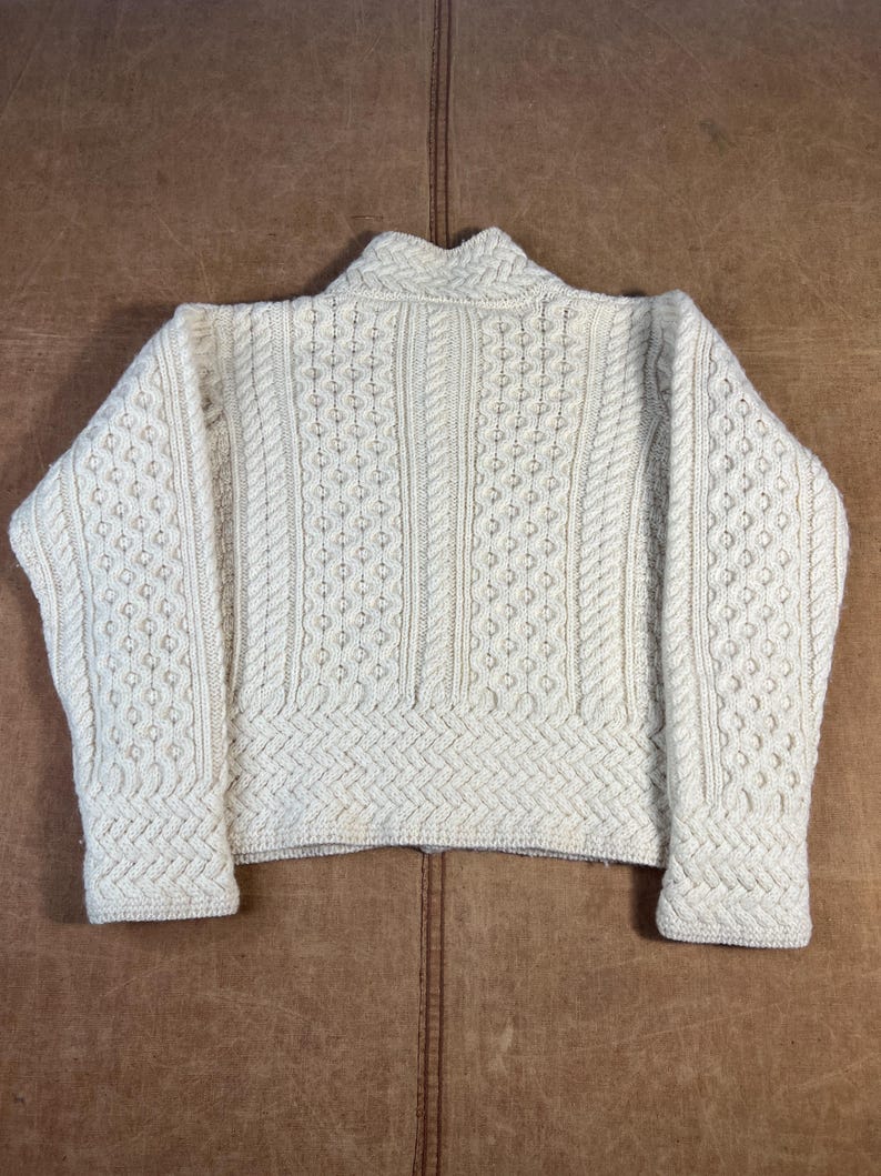 May include: Cream-colored, textured knit sweater with a high collar. The sweater features intricate cable knit and diamond patterns across the body and sleeves. The garment is laid flat on a brown leather surface.