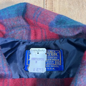 May include: Close-up of a red, blue, and teal plaid wool shirt collar. The shirt has a blue lining and a tag that reads "Pendleton" and "Virgin Wool U.S.A.". The tag also has the text "Metric Sports".