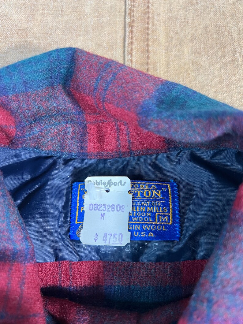 May include: Close-up of a red, blue, and teal plaid wool shirt collar. The shirt has a blue lining and a tag that reads "Pendleton" and "Virgin Wool U.S.A.". The tag also has the text "Metric Sports".