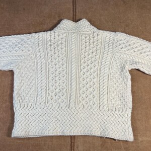 May include: Cream-colored, cable-knit sweater with a high collar. The sweater features various textured patterns, including cable and diamond designs. The garment is made of a thick, warm material, perfect for cold weather. The sweater is laid flat on a brown surface.