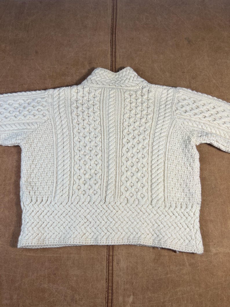 May include: Cream-colored, cable-knit sweater with a high collar. The sweater features various textured patterns, including cable and diamond designs. The garment is made of a thick, warm material, perfect for cold weather. The sweater is laid flat on a brown surface.