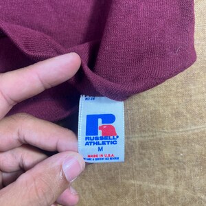 May include: Close-up of a maroon-colored garment with a tag. The tag is white with blue and red lettering, displaying the brand "RUSSELL ATHLETIC" and the size "M". The tag also indicates the item was made in the U.S.A.