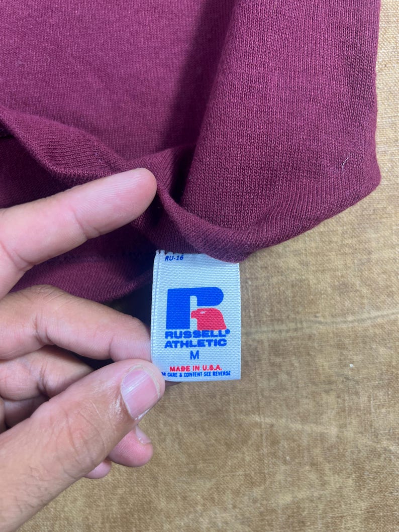 May include: Close-up of a maroon-colored garment with a tag. The tag is white with blue and red lettering, displaying the brand "RUSSELL ATHLETIC" and the size "M". The tag also indicates the item was made in the U.S.A.